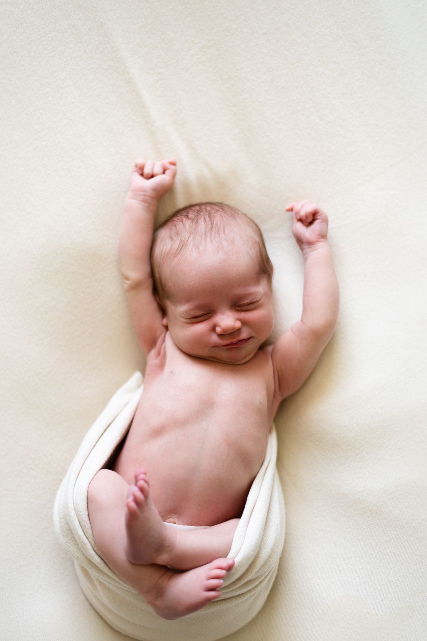 Happy newborn stretching with arms up while partially wrapped in white fabric.