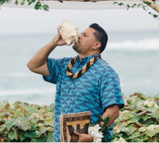 A traditional Hawaiian officiant blowing into a conch shell during a ceremony by the ocean on Maui, Hawaii.