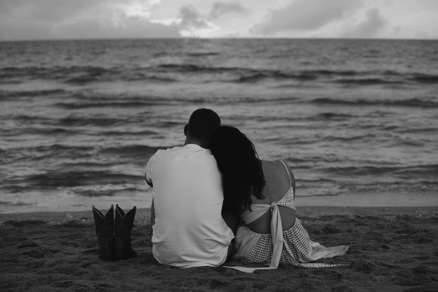 Two people sit together on a beach at dusk in black and white, watching the waves roll in.