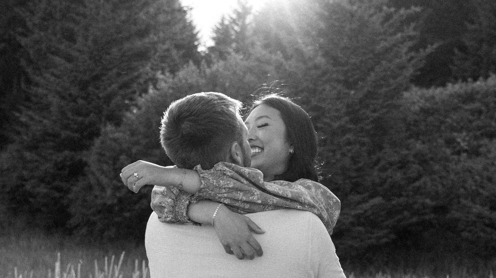 A joyful couple embraces in a sunlit forest setting, captured in black and white photography.