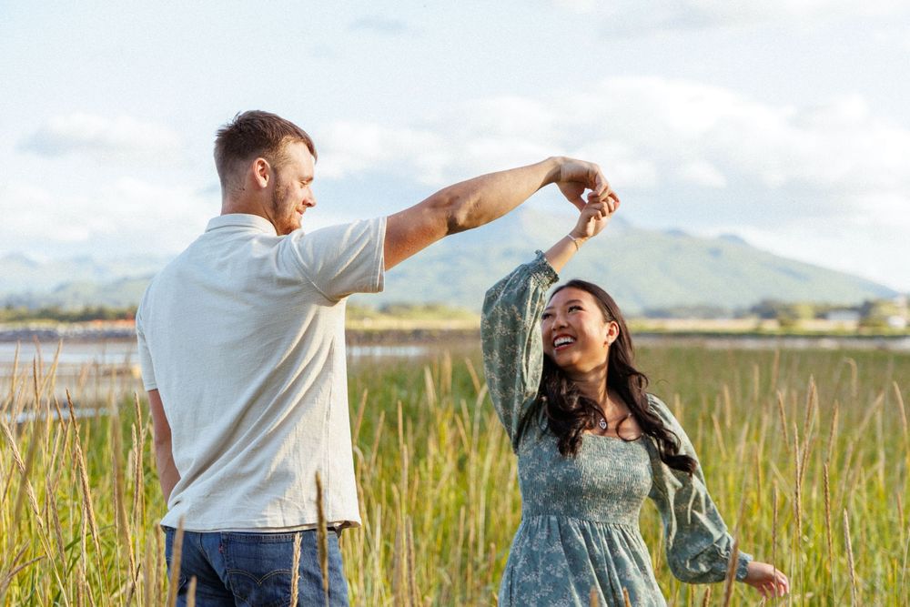 A couple dances together in a grassy field on a sunny day with mountains visible in the distance.