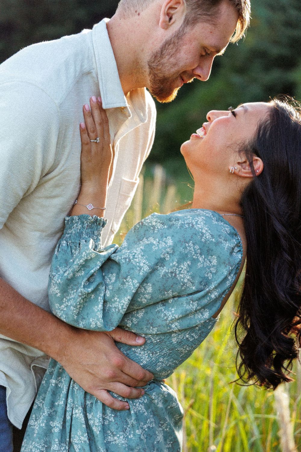 Romantic couple embraces in a field wearing white shirt and blue floral dress during sunset photoshoot.