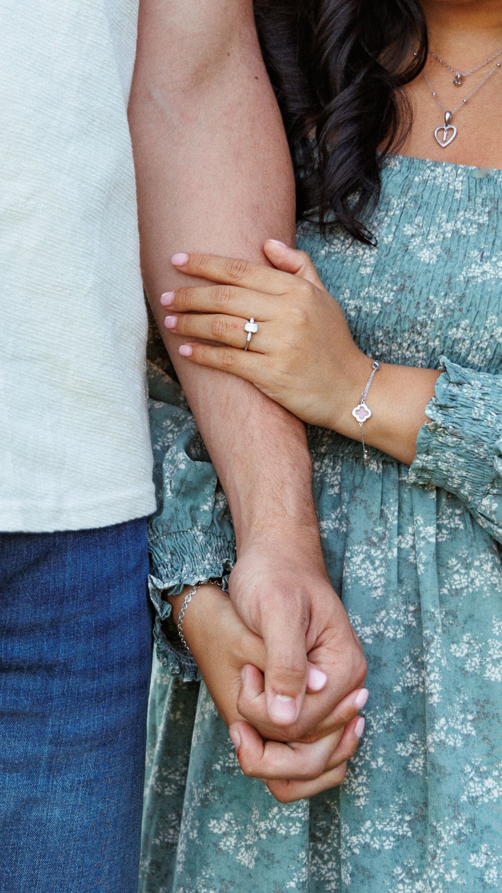 Close-up of couple holding hands wearing engagement ring, woman in floral turquoise dress with heart necklace.