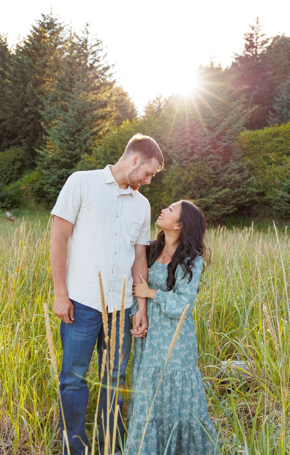 A romantic couple stands together in a sunlit field of tall grass with evergreen trees in the background.
