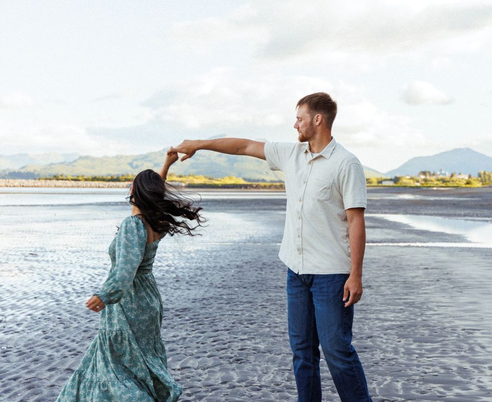 Two figures in casual attire dance together at sunset on a beach with mountains in the background.