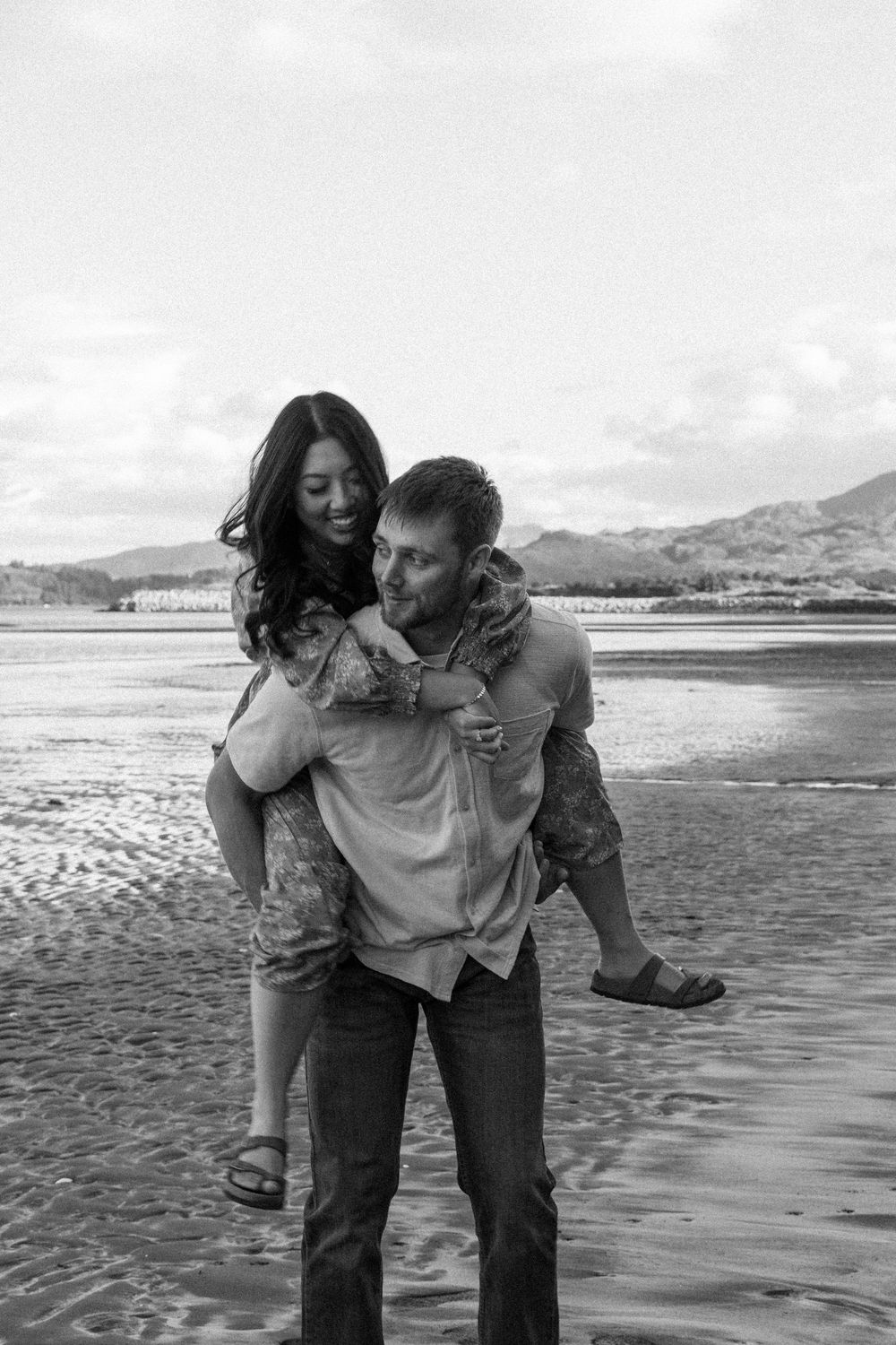 A joyful piggyback ride on the beach with mountains in the background captured in black and white.