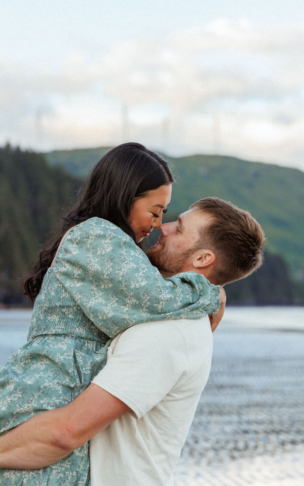 A couple sharing a tender romantic moment by the water with mountains and forest in the background.