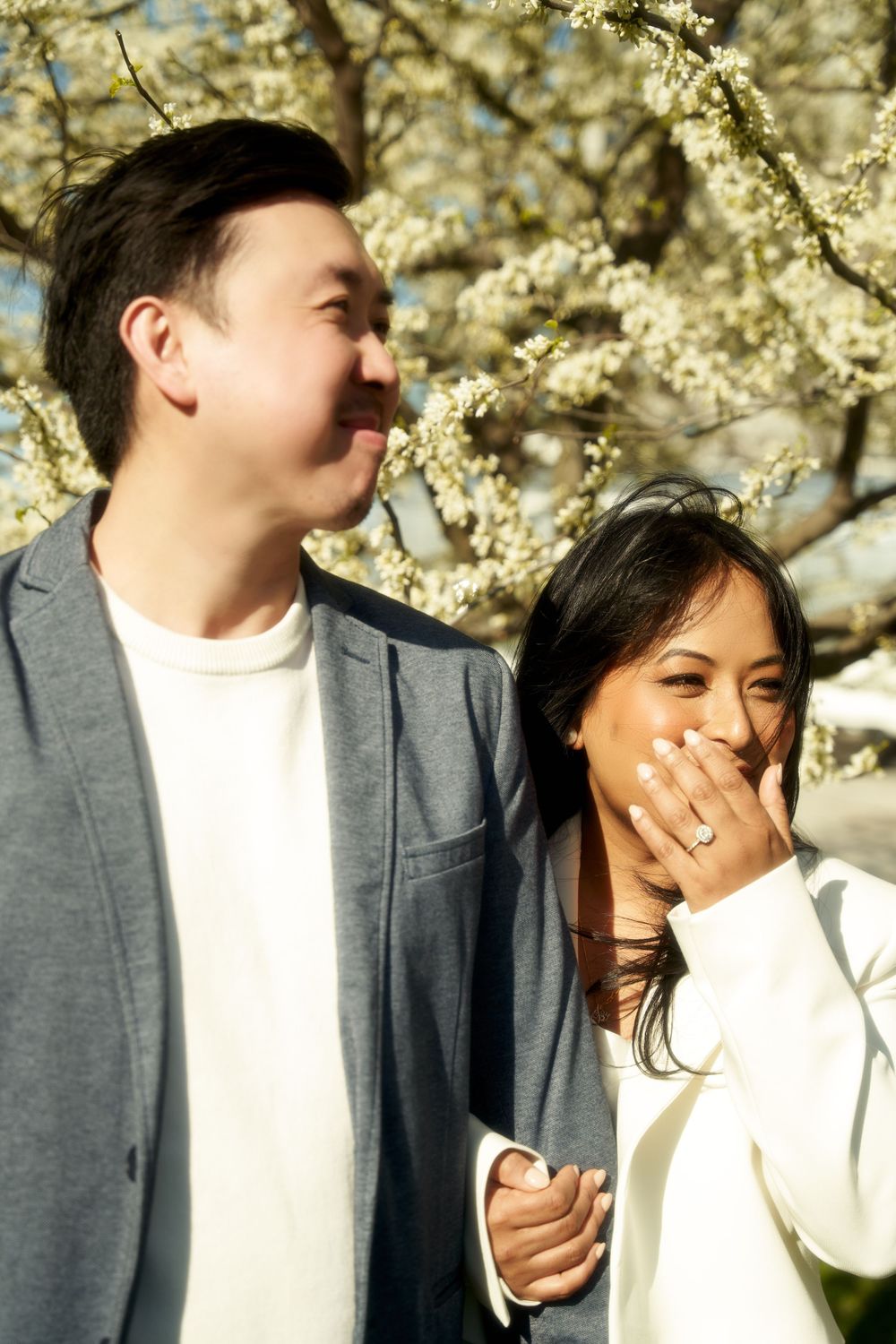 A couple shares joyful laughter together against a backdrop of blooming cherry blossoms on a sunny spring day. Engagement  in Williamsburge