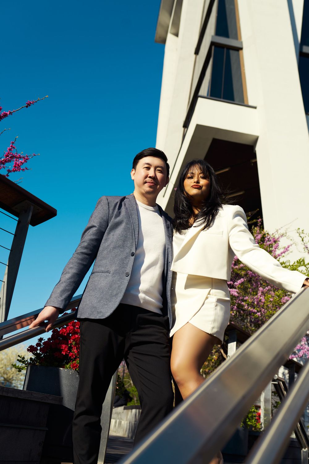 Couple walking along a white architectural exterior with vibrant pink flowers against bright blue sky. Engagement  in Williamsburge