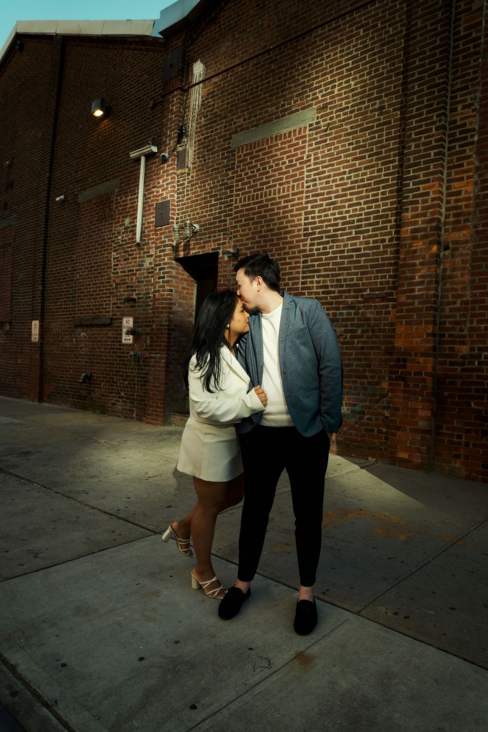 Couple strolling along an urban brick wall in dramatic lighting creating long shadows on the pavement. Engagement  in Williamsburge