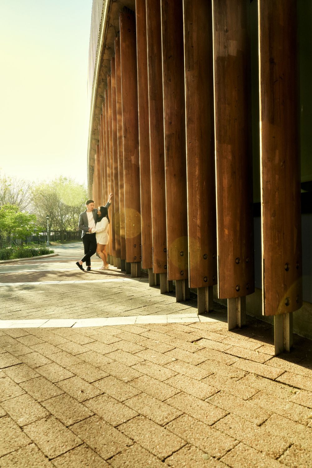 Two people stand between tall wooden pillars along a brick path on a sunny day in an architectural setting. Engagement  in Williamsburge