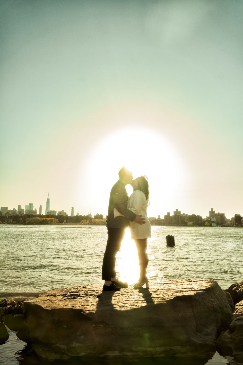 Silhouetted couple stands together on rocky shore against bright glowing sunset with city skyline in distance. Engagement  in Williamsburge