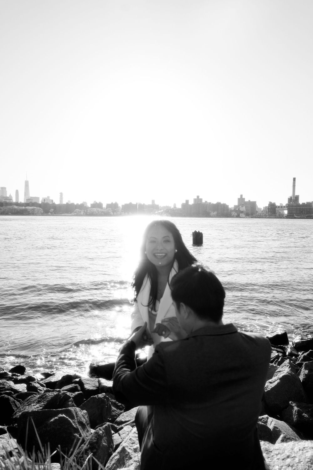 A black and white photo of two people sitting on rocky shoreline with city skyline visible across the water. Engagement  in Williamsburge