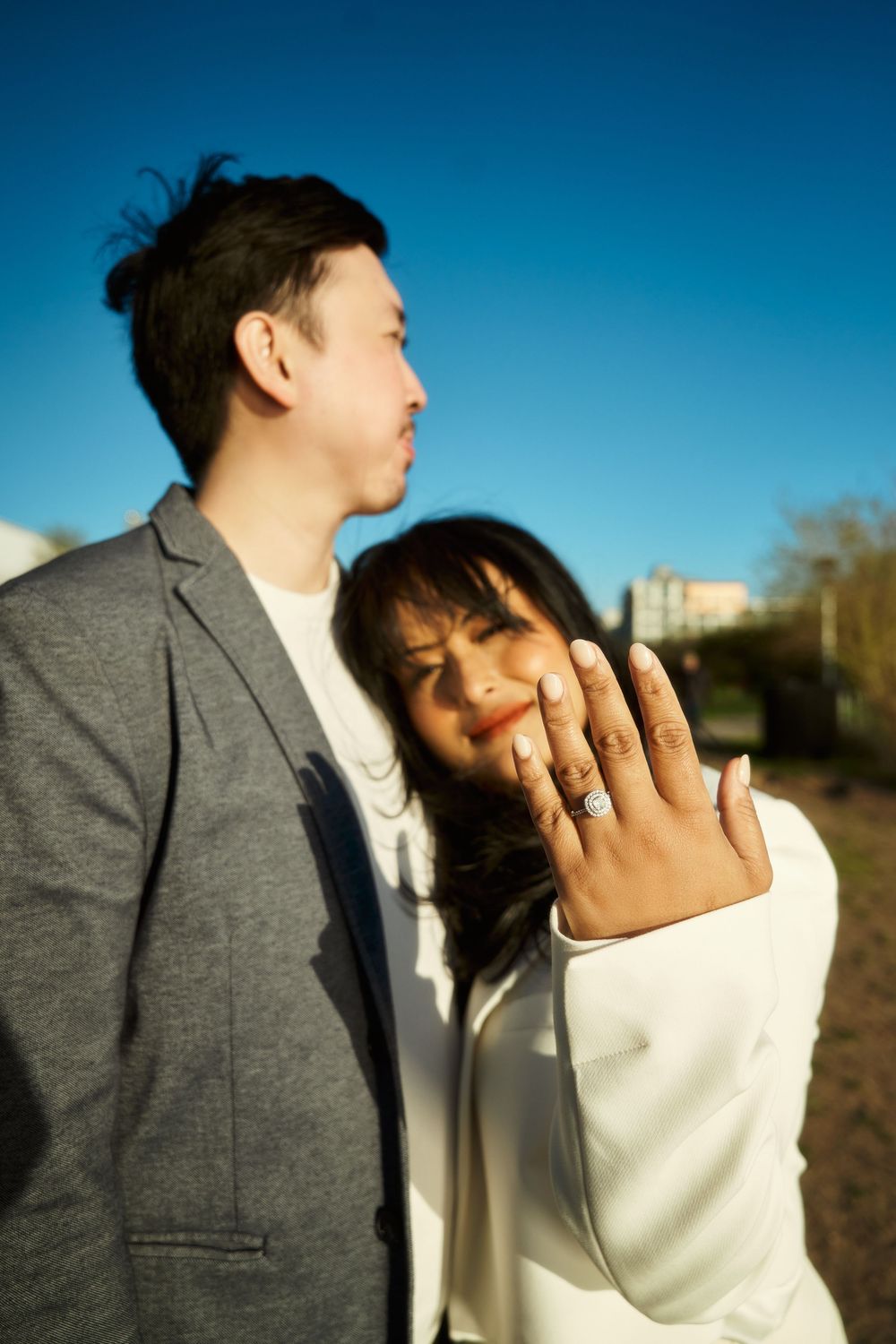 A couple wearing white and gray formal attire shares an intimate moment against a blue sky backdrop. Engagement  in Williamsburge