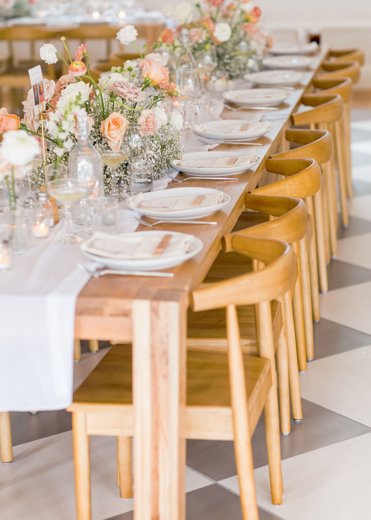 Long wooden dining tables set with white linens, glassware, and floral centerpieces paired with elegant tan dining chairs.