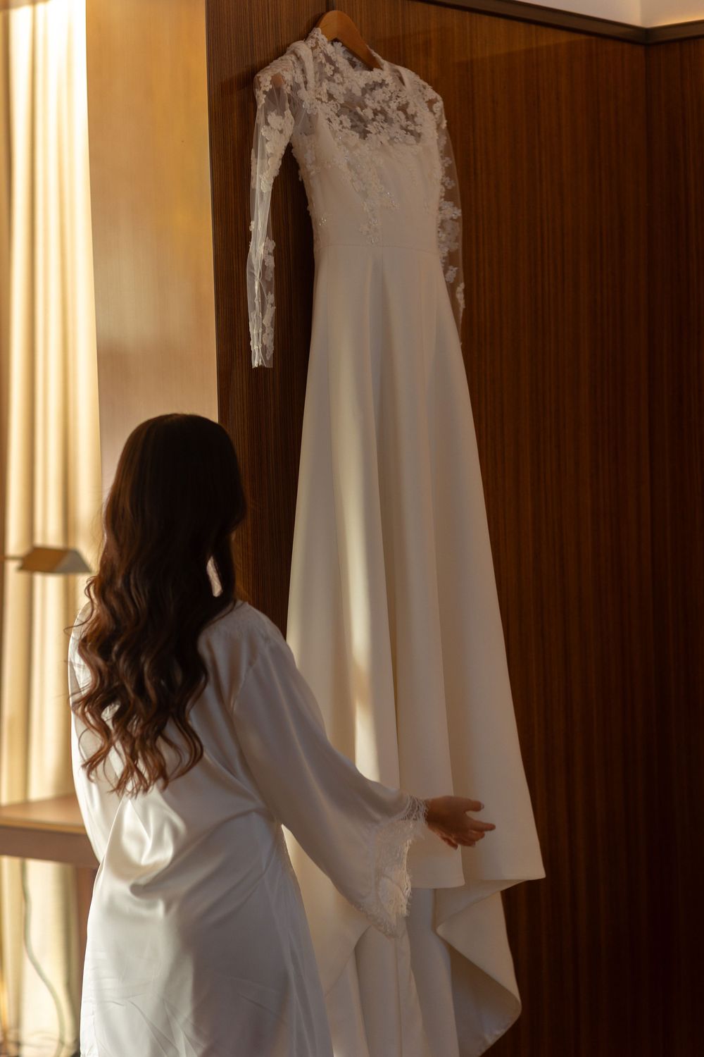 Person in white robe reaching toward elegant white wedding dresses hanging by window in sunlit dressing room.