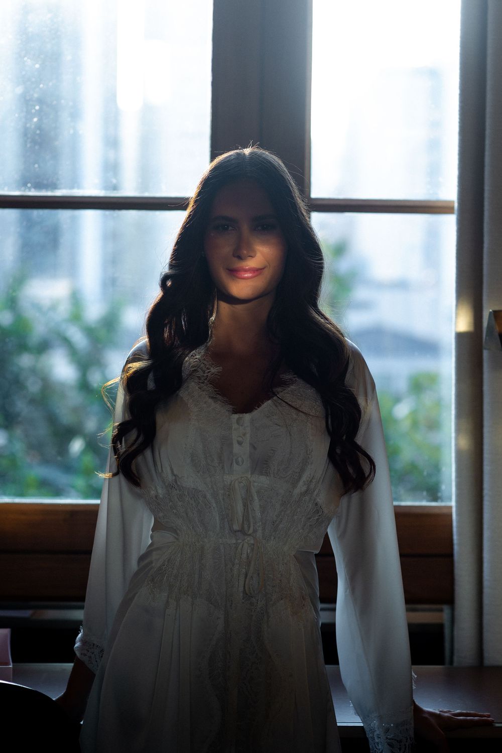 A silhouetted figure in a white dress stands by a window in a dark room creating a moody atmospheric scene.