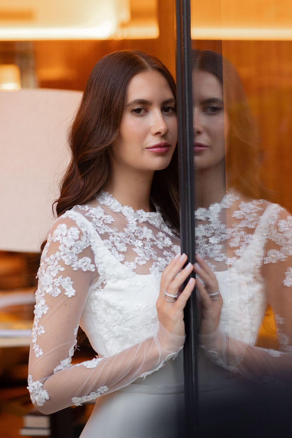 Glamorous bride in long sleeve white lace dress poses elegantly by window with warm lighting.