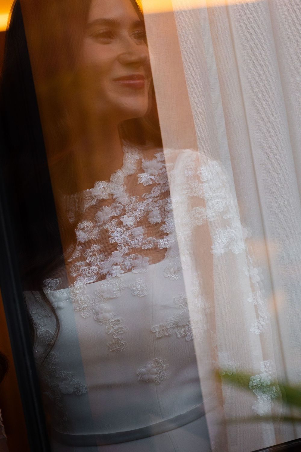 Woman in white lace dress gazes through sheer curtains at golden sunset creating dreamy atmosphere.