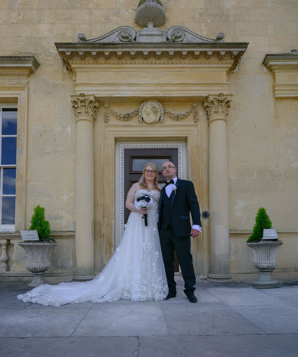 Wedding couple stands in front of elegant stone building with classical architecture and decorative urns.