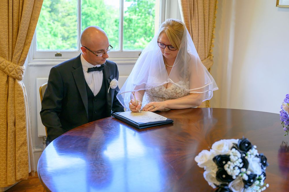 Newlyweds signing their marriage documents at an elegant table with yellow curtains.