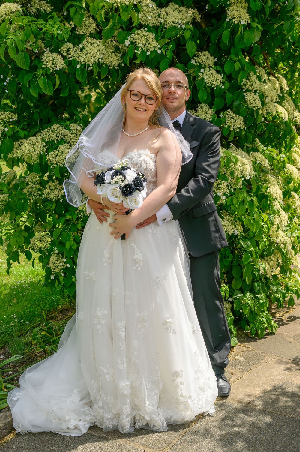 Bride and groom pose lovingly in a garden setting with blooming white flowers and lush greenery.
