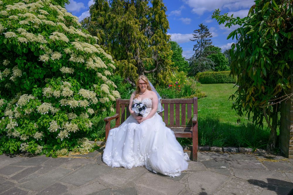 Couple in wedding attire poses in a lush garden setting with white floral blooms and greenery on a sunny day.
