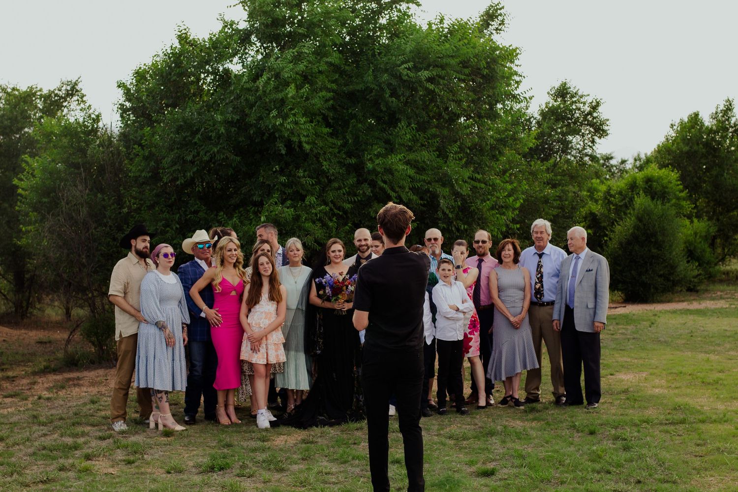 Wedding party gathers for outdoor group photos in lush green garden setting during summer ceremony.