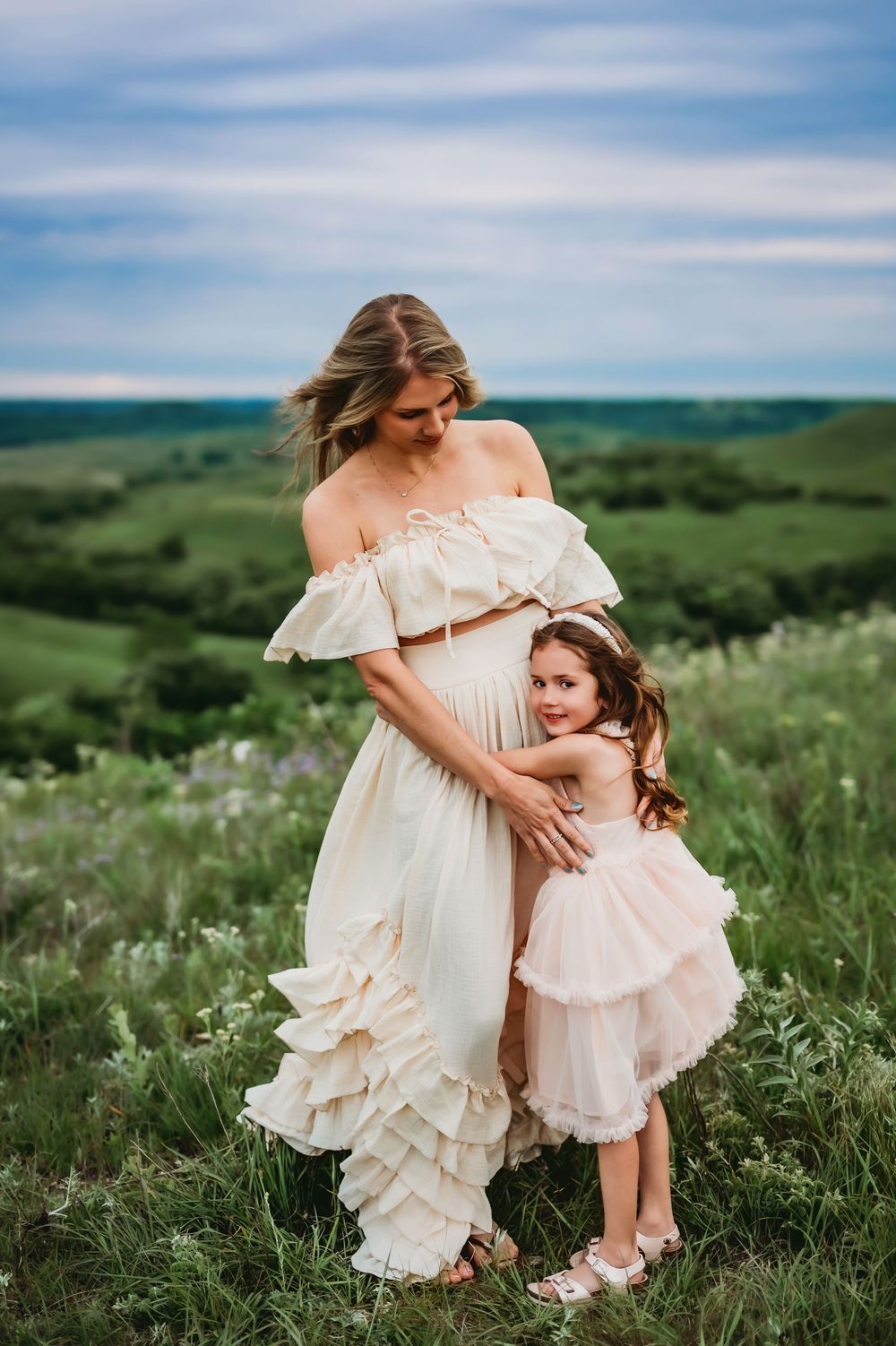 Two people in flowing cream and pink dresses embrace while standing in a scenic green meadow under blue skies.