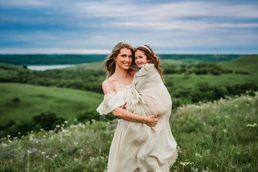 Two people in flowy white dresses embrace while standing in a scenic green meadow with wildflowers on a sunny day.