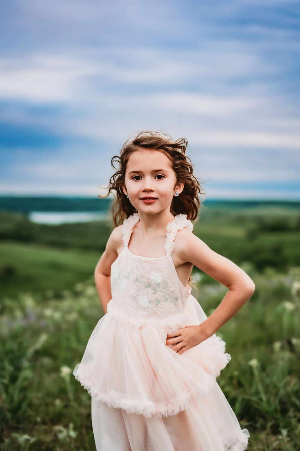 A child wearing a light pink lace dress twirls in a green meadow under blue skies.