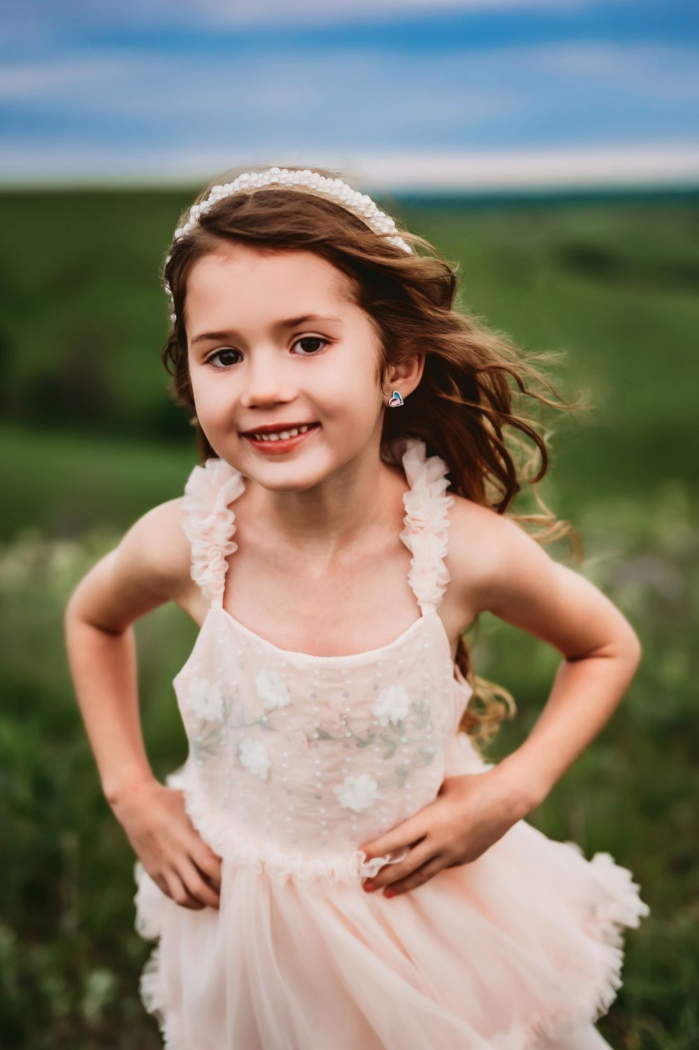 A child in a light pink ruffled dress poses playfully outdoors against a green natural backdrop.