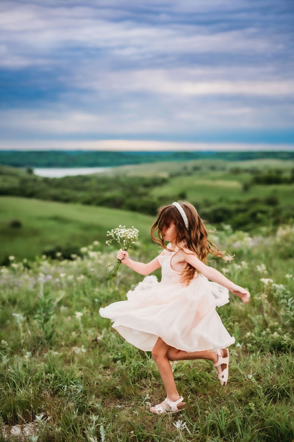 A girl in a flowing pink dress spins joyfully in a meadow of wildflowers under cloudy summer skies.