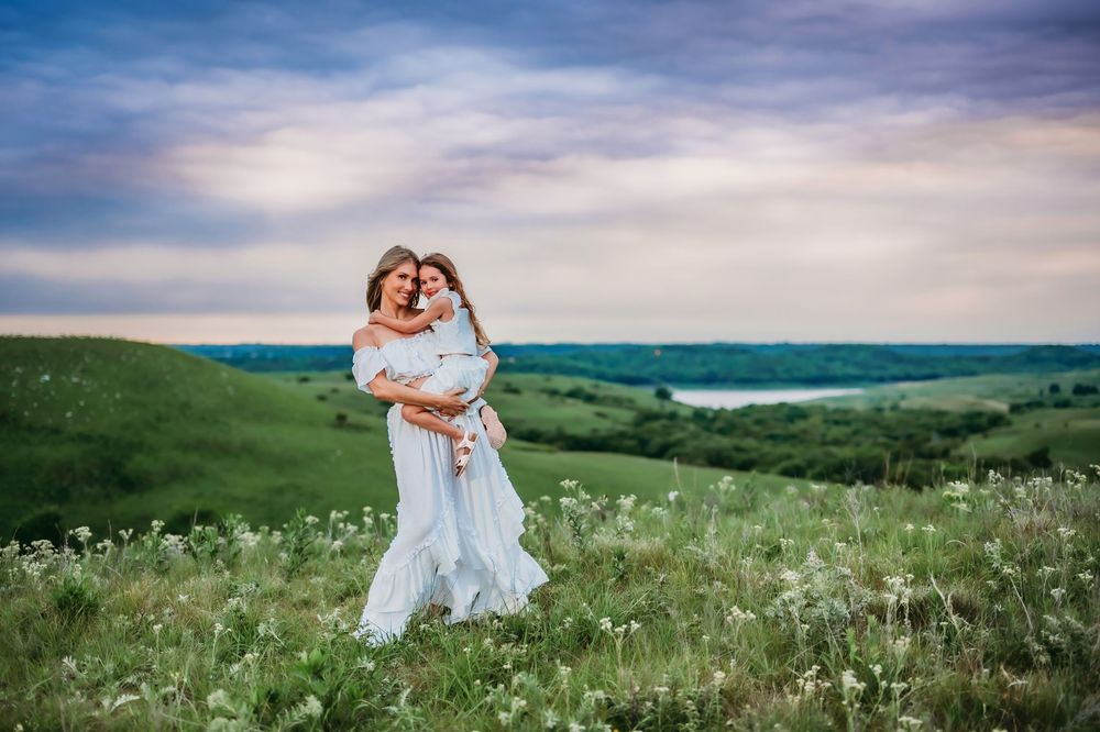 Couple embracing in a sweeping green meadow with white wildflowers under a dramatic cloudy sky.