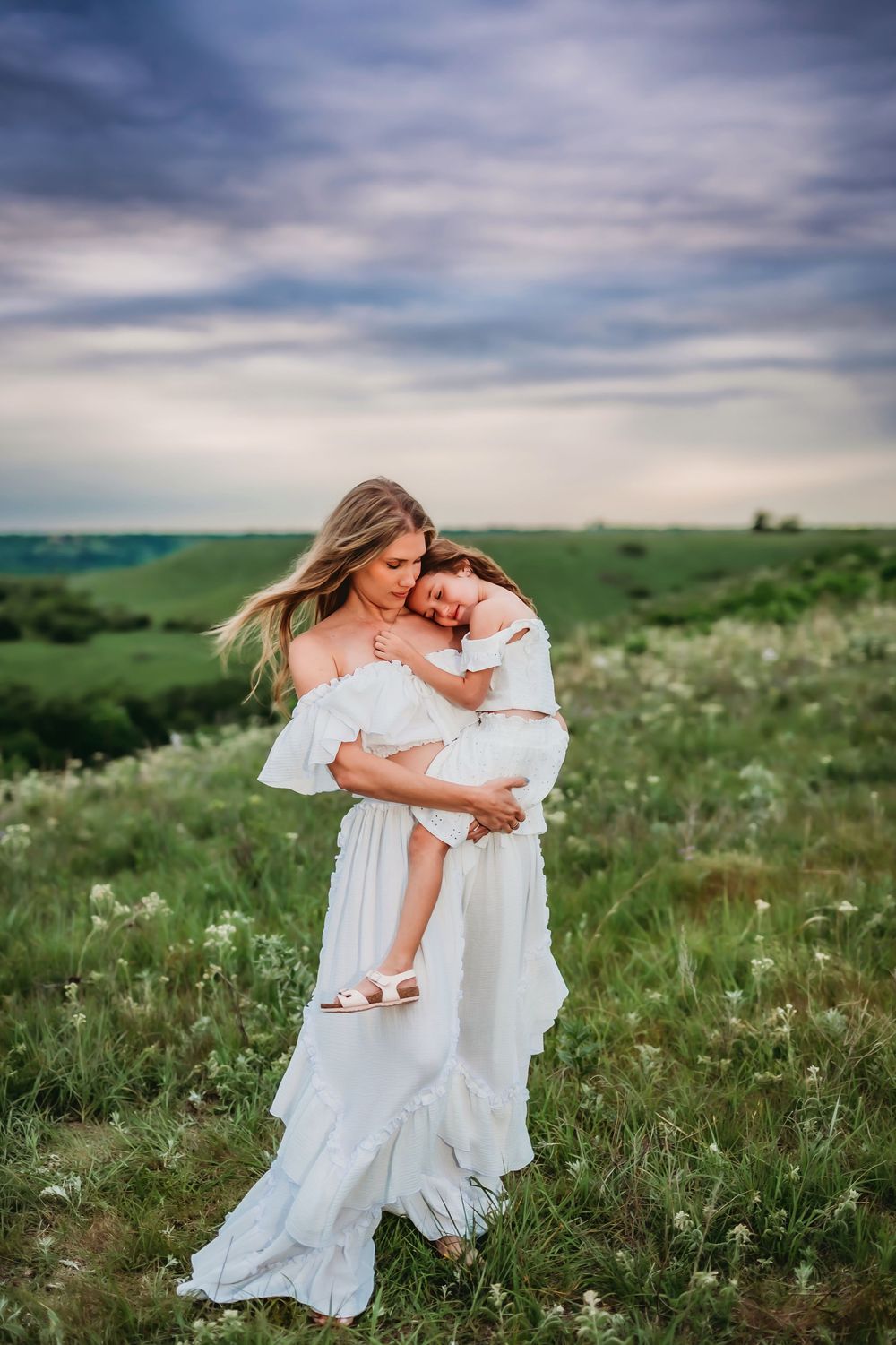 Mother and daughter in flowing white dresses embrace tenderly in a wildflower meadow under cloudy skies.