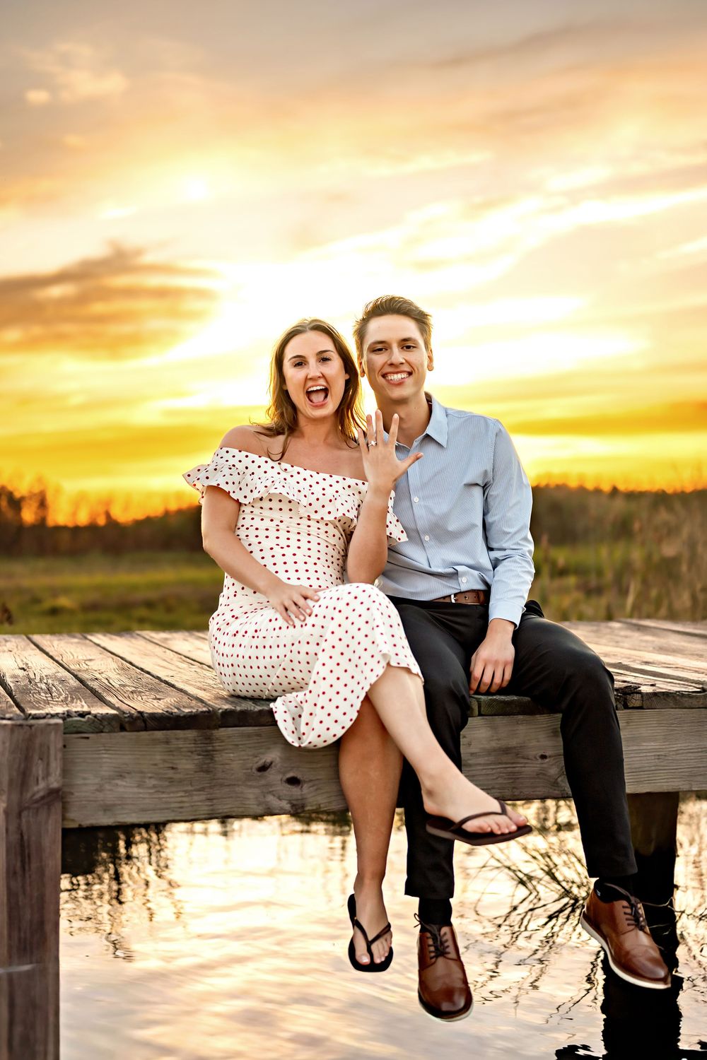 A couple sits playfully on a wooden dock together during a vibrant golden sunset over a lake.