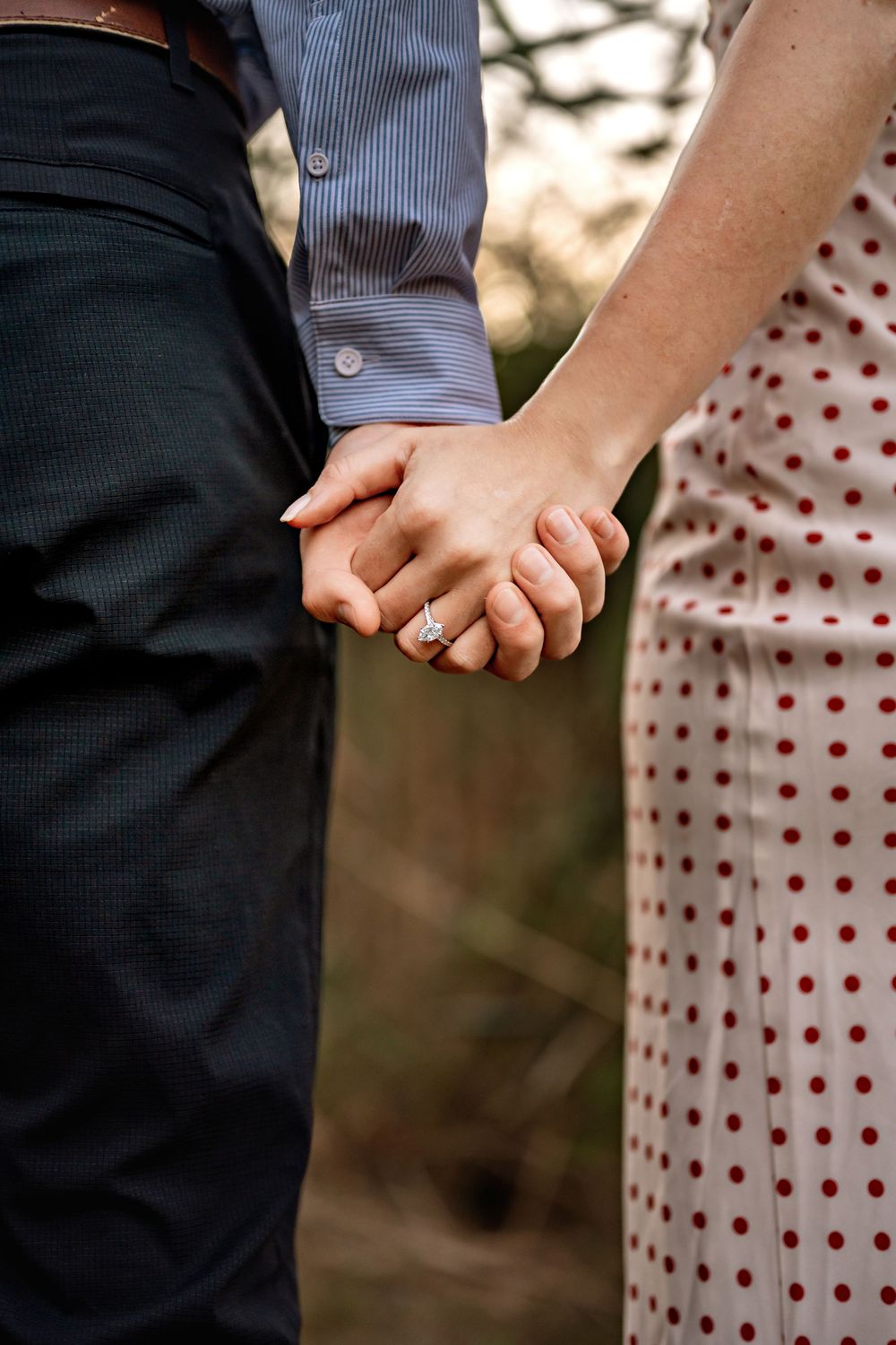 Close-up of two people holding hands with engagement ring visible, wearing polka dot dress and dark pants.