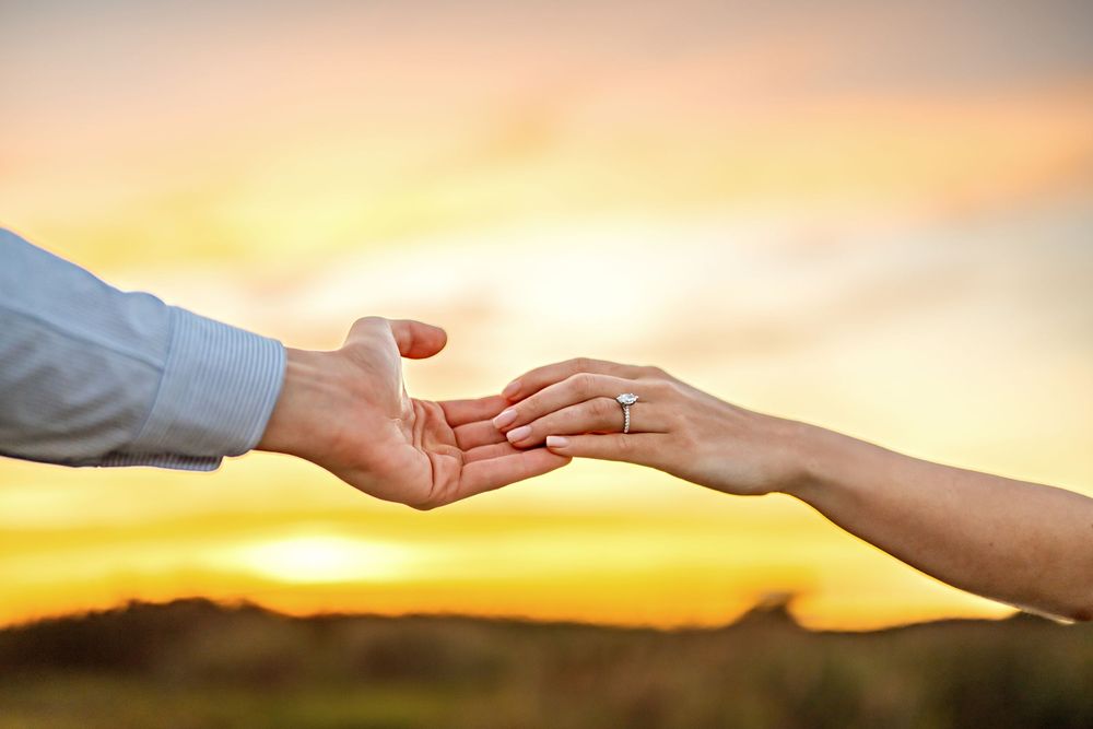 Two hands reaching out to connect against a warm sunset sky with soft clouds in the background.