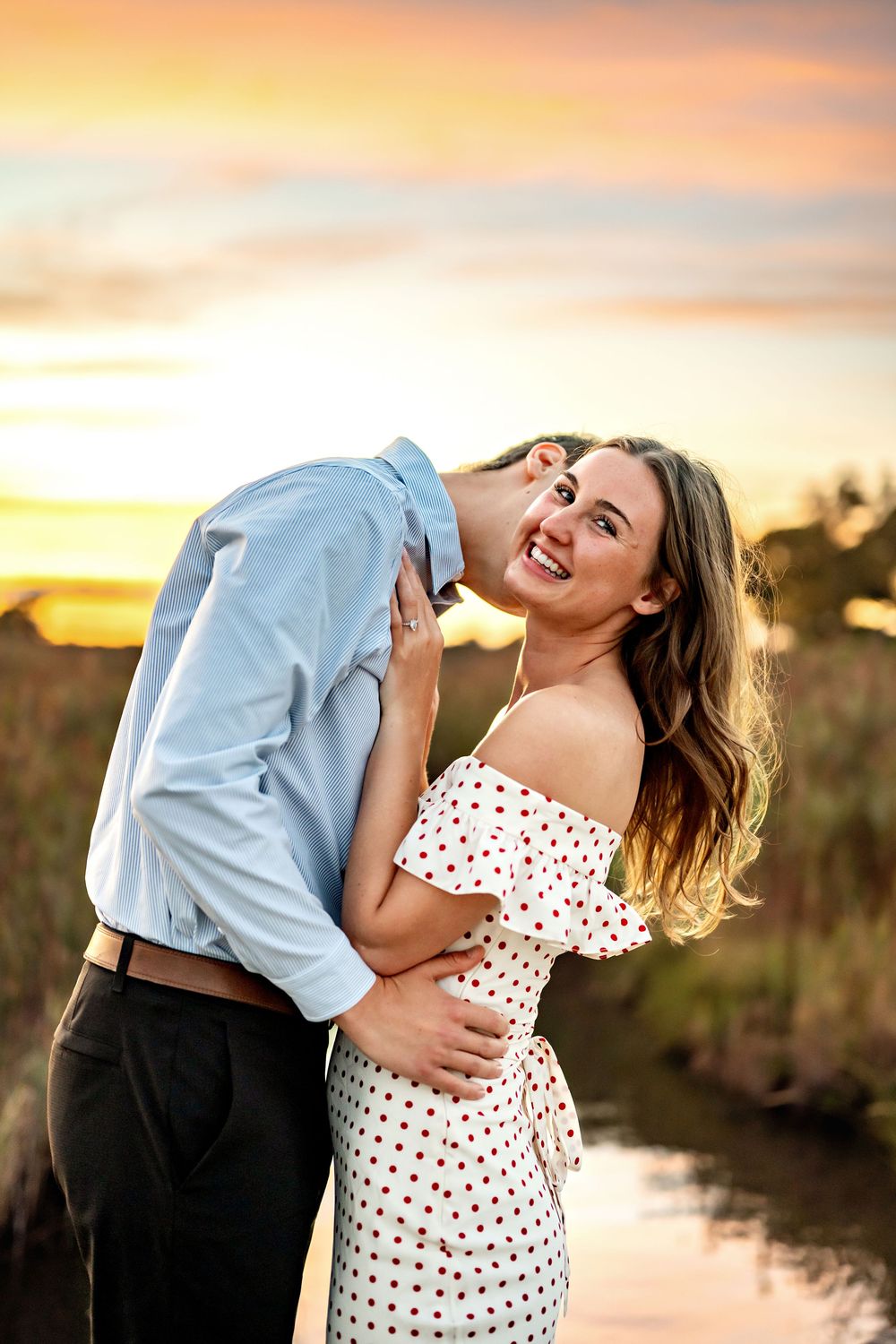 A couple embraces at sunset by the water as the sky glows with orange and pink hues.