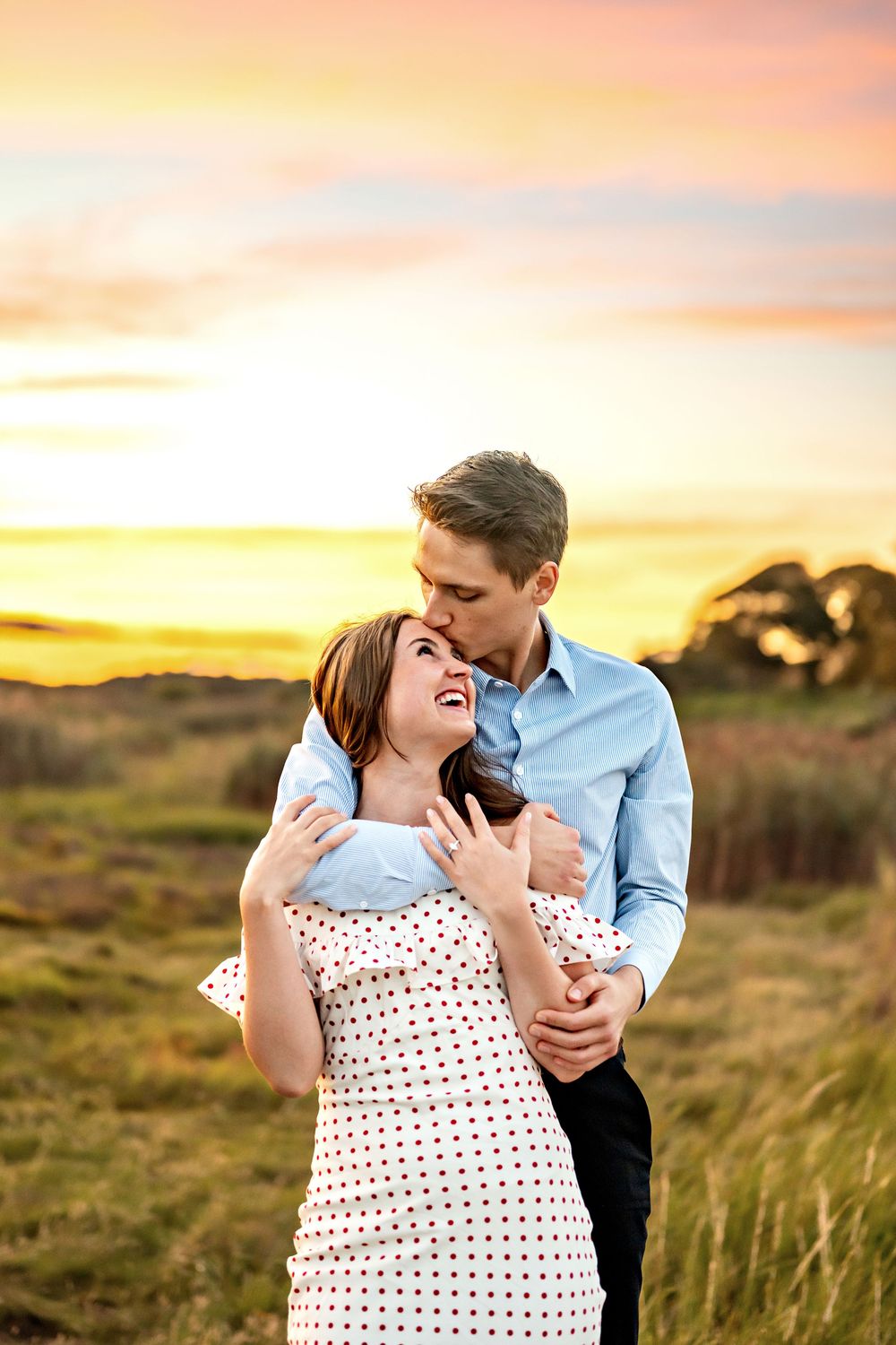 A couple embraces romantically in a grassy field during a golden sunset.