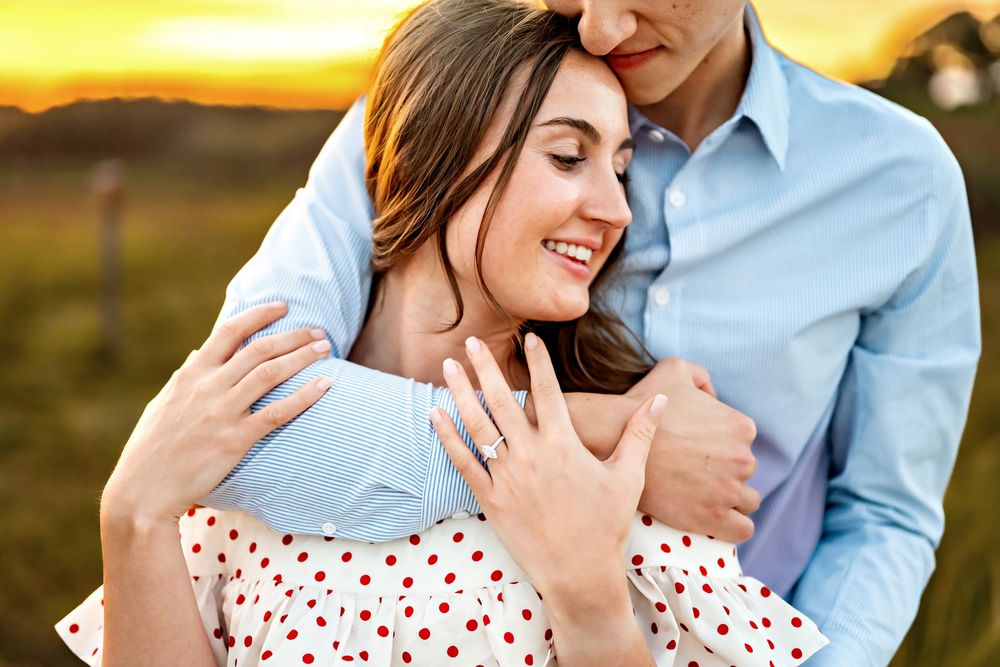 Couple in romantic embrace at sunset wearing blue shirt and polka dot dress showing engagement ring.
