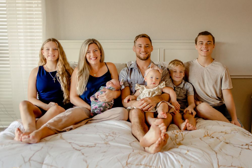 A family sits together on a white bed in warm lighting wearing casual clothes for in-home session in Keller, TX