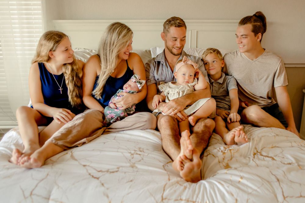 A family sits together on a white bed in warm lighting wearing casual clothes for in-home session in Keller, TX