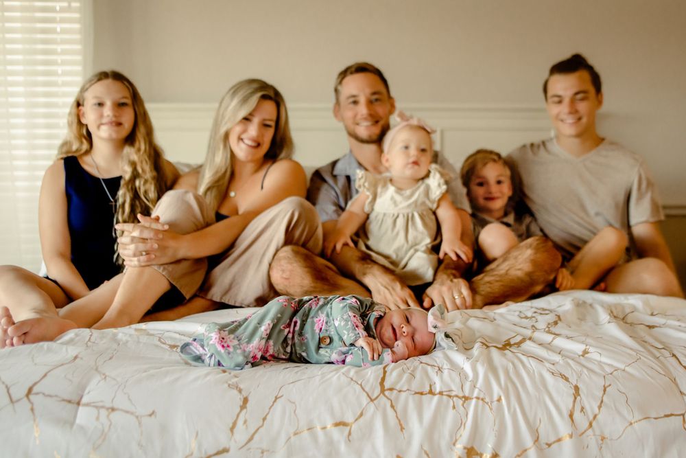 A family sits together on a white bed in warm lighting wearing casual clothes for in-home session in Keller, TX
