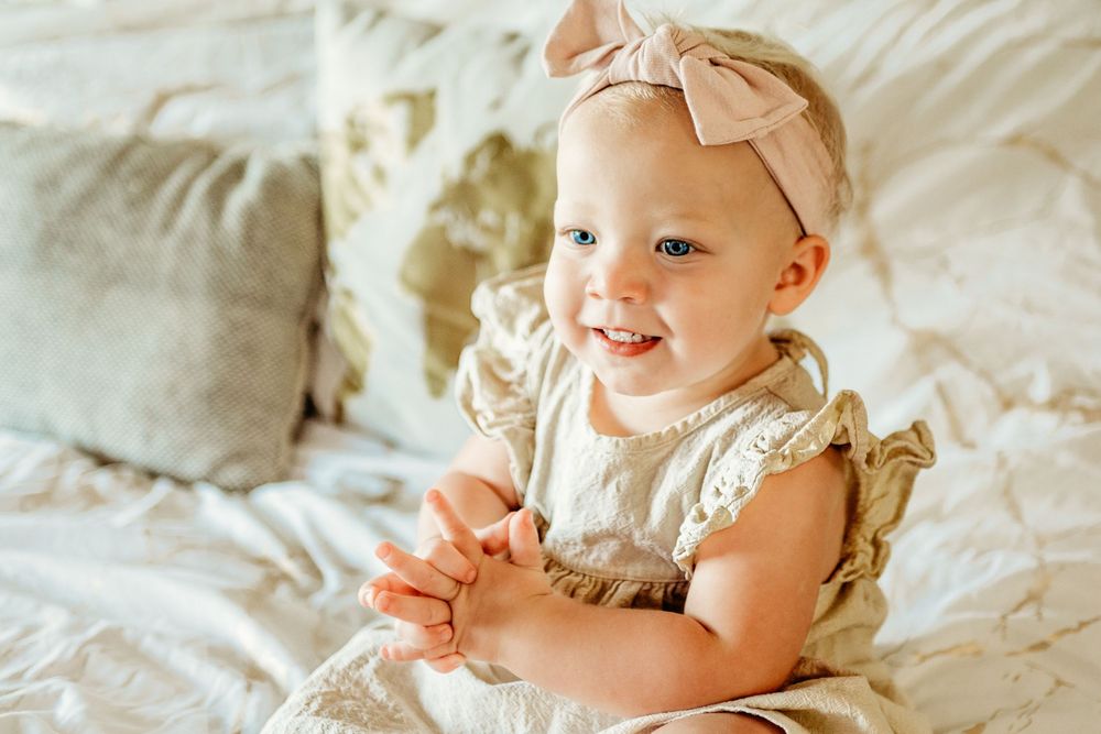 Smiling baby wearing a beige lace dress and pink bow headband sits on white bedding in fort worth in-home photo session