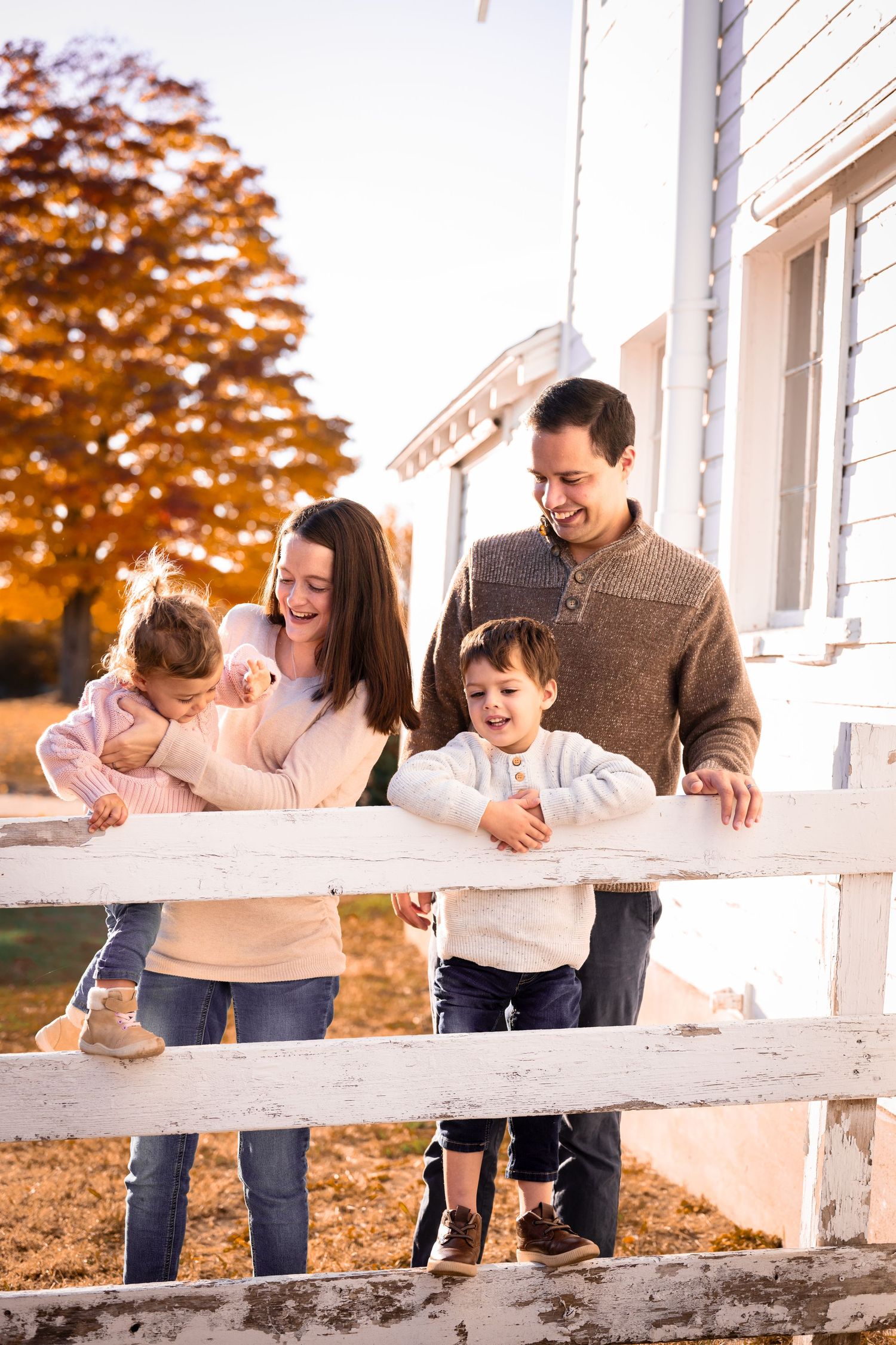 Joyful family of four standing by a white fence outside a farmhouse during golden autumn season.