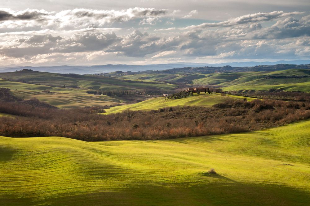Rolling green hills stretch across the scenic Tuscan countryside under dramatic clouds and sunlight.