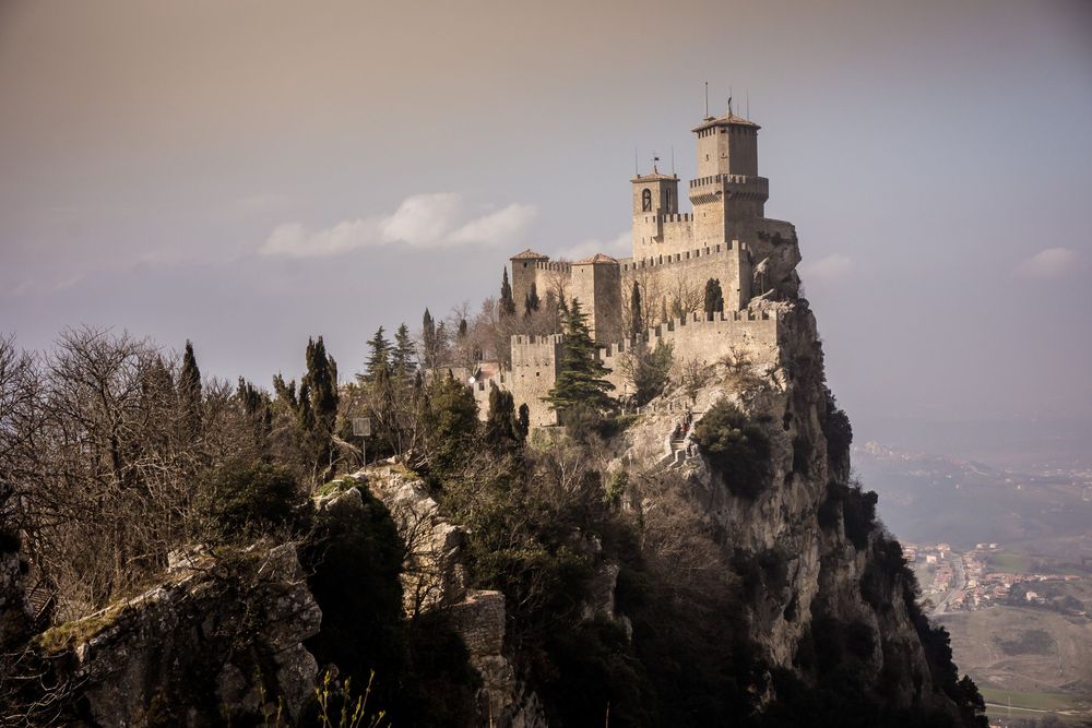 Medieval stone castle fortress atop a rocky mountain ridge overlooking a misty winter landscape.