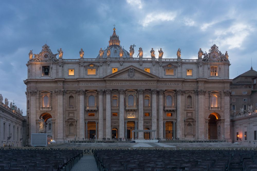 Panoramic view of St. Peter's Basilica illuminated at dusk in Vatican City with dramatic cloudy sky.