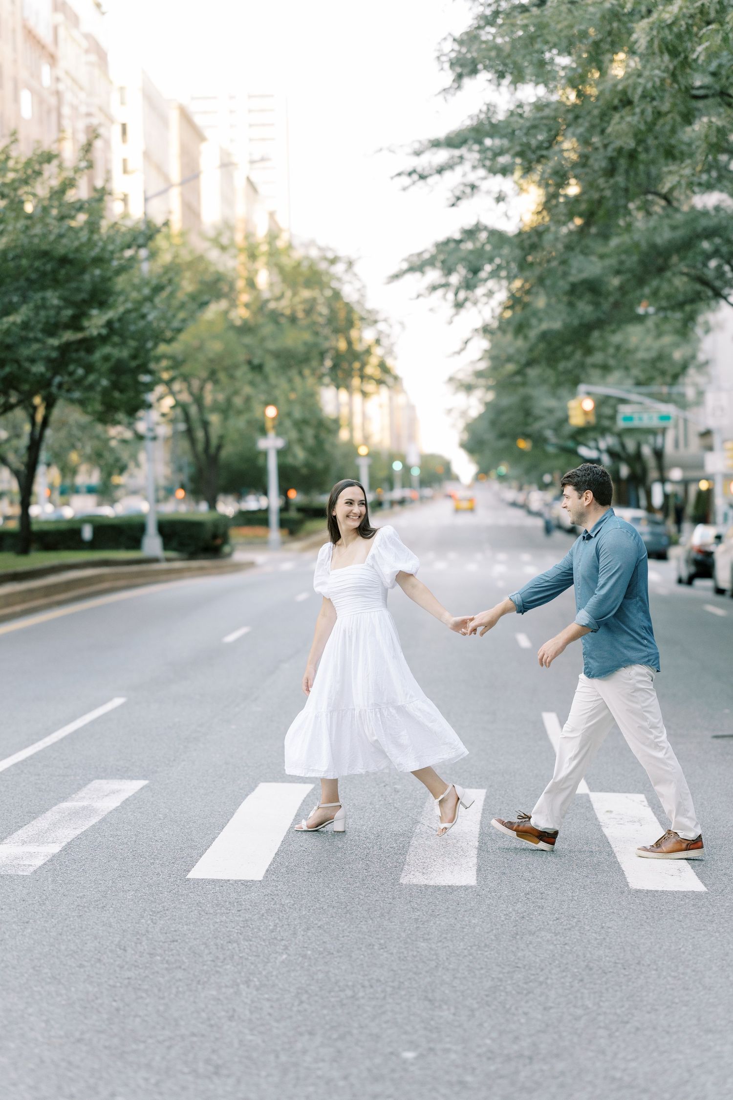 A couple playfully crosses a crosswalk in an urban setting, recreating the iconic Beatles Abbey Road album cover.